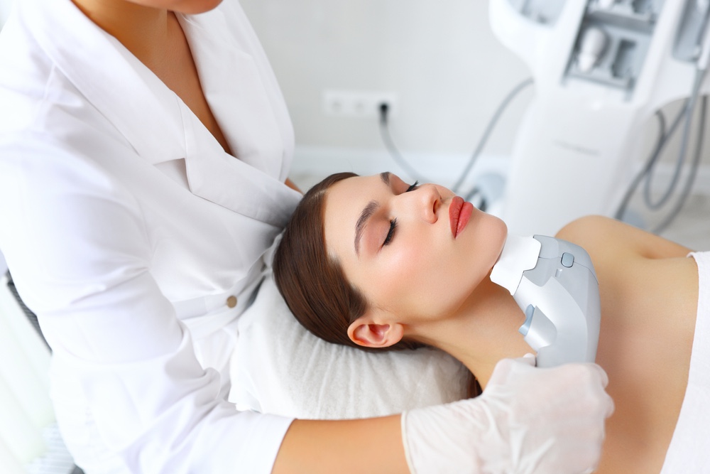 HiFu treatment | A white woman laying on a treatment chair. Her aesthetician is behind her and is treating her jawline area with the HiFu machine.